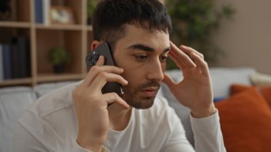 Akıllı telefondan konuşan genç adam endişeli bir ifadeyle modern oturma odasında oturuyor.