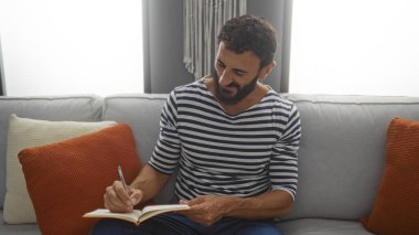 Çizgili tişörtlü, sakallı genç adam evdeki kanepede turuncu ve beyaz yastıklarla otururken not defterine yazıyor.