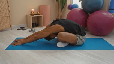 Spor salonundaki mavi yoga minderinin üzerinde uzanan genç bir adam. Arka planda mumlar, halterler ve egzersiz topları var.