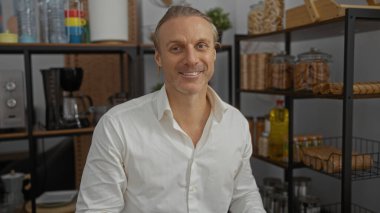 Beyaz tişörtlü, sarı saçlı orta yaşlı bir adam. Ofis ortamında gülümsüyor. Arka planda raflar ve ev eşyaları var..