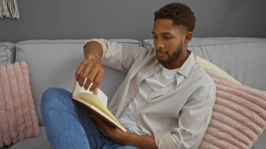Genç adam modern dairenin oturma odasındaki koltukta kitap okuyor.