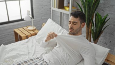 Yatak odasındaki yakışıklı genç adam kendini bir gömlekle yelpazeliyor, aşırı ısınmış görünüyor, şık, modern, kapalı bir ortamda doğal ışık altında bir yatakta yatıyor..