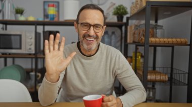 Ofis ortamında gülümseyen bir adam elinde kupayla arka planda modern raflarla kendine güvenerek selam veriyor..