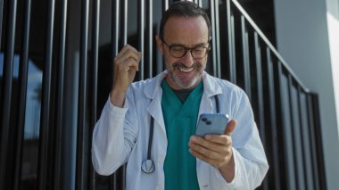 Tıbbi kıyafetli İspanyol bir adam, şehir merkezinde bir hastanenin önünde dururken akıllı telefondan iyi haberleri kutluyor..