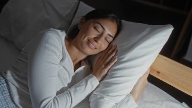 Yüzünde bir gülümseme, esmer saçlarını ve İspanyol özelliklerini vurgulayarak, rahat bir yatak odasında huzur içinde uyuyan bir kadın.