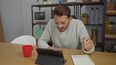 Genç adam ofisinde tablet ve not defteriyle çalışıyor, gözlük ve süveter giyiyor, kırmızı bardakla oturmuş, mikrodalga ve şişelerle çevrili.