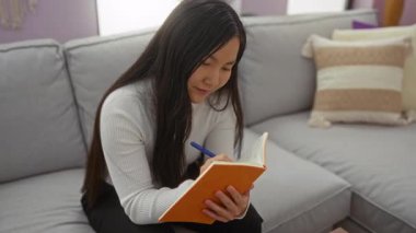 Evdeki kanepede, beyaz kazak giyen, uzun saçlı, parlak oturma odası ortamında kapalı, odaklanmış ve rahat görünen genç bir kadın..