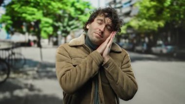 Kapalı gözlü genç adam yeşil ağaçlar ve şehir arka planına sahip bir caddede dinleniyor, kahverengi bir ceket giyiyor ve rahat ve hoşnut görünüyor..