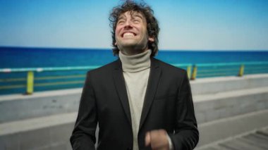 Genç bir adam canlı mavi bir okyanusun önünde kumsalda gülümsüyor ve pozitiflik saçan büyüleyici bir açık hava portresi içinde keyif ve rahatlama hissi veriyor..