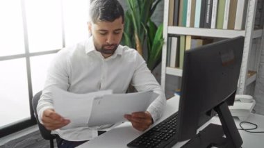 Parlak bir ofiste çalışan genç adam masasında, etrafı kitaplarla dolu bir monitör, evrak ve raflarla çevrili belgeleri inceliyor..