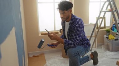 Genç adam yeni oturma odasının duvarını boyuyor, boya silindiriyle ve projede rehberlik için telefonu kullanıyor..