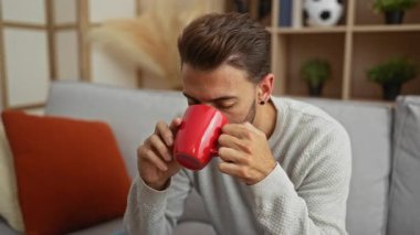 Genç adam, modern dekor ve kırmızı bardakla dekore edilmiş bir oturma odasında rahat bir koltukta oturup kahveyi seviyor..