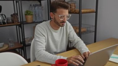 Modern bir ofis ortamında çalışan genç bir adam dizüstü bilgisayar ve kırmızı kahve fincanı ile ahşap masada odaklanma ve profesyonellik gösteriyor..