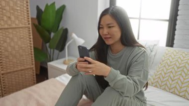 Yatakta rahat bir yatak odasında oturan bir kadın akıllı telefon kullanıyor, ifadelerini sergiliyor, yeşil bitkiler ve yumuşak ışıklandırma ile modern iç mekanı vurguluyor..