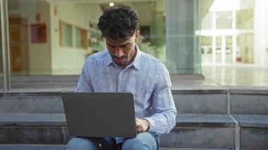 Şehir merdivenlerinde dizüstü bilgisayarda çalışan genç adam şehir ortamında düşünceli görünüyor..