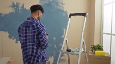 Genç adam modern oturma odası düzeninde referans olarak akıllı telefon kullanarak duvarları boyuyor..