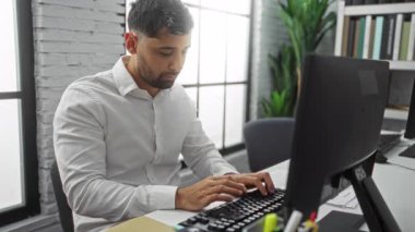 Ofisteki bir masada çalışan, bilgisayar kullanan, modern ve kapalı bir ortamda üretkenlik odaklı genç bir adam..