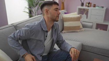 Modern bir oturma odasında koltukta otururken sırt ağrısı çeken genç bir adam, yumuşak ışıklandırma ve şık dekorasyon unsurlarıyla çağdaş bir iç mekan sergiliyor..