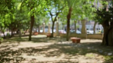 Bulanık açık hava parkı sahnesi, yeşil ağaçlar ve çözülmüş arka plan. Bokeh efektleriyle güneşli bir ortam yakalıyor..