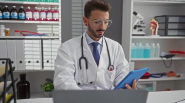 Ensesinde steteskopla klinikteki genç İspanyol erkek doktor tıbbi malzeme ve raflarla çevrili dijital tablet üzerinde yoğunlaşıyor..