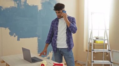 Oturma odasında ev tadilatı yapan genç bir adam. Dizüstü bilgisayar ve telefonla gündelik kıyafetleri ve modern iç dekorasyonları gösteriyor..