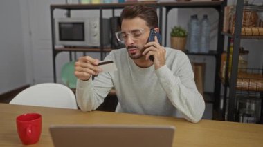 Ofisteki genç adam elinde kredi kartıyla telefonla konuşuyor, gözlük takıyor ve kırmızı bardaklı bir dizüstü bilgisayarla masada oturuyor..