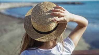 Hasır şapkalı kadın güneşli sahil gezintisinde deniz manzarasının keyfini çıkarıyor, beyaz gömlek ve saat giyiyor, yazın deniz kenarında uzun saçlarıyla sergileniyor..