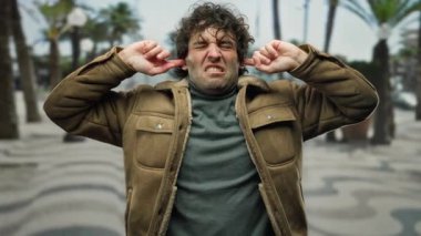 Şehirdeki açık havada, kahverengi ceketli, kıvırcık saçlı, gözleri kapalı, kulakları tıkayan, hayal kırıklığını ifade eden bir adam gündüz vakti bulanık bir palmiye ağacında....