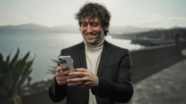 Genç adam, güneşli bir sahil kenarında akıllı telefon kullanırken gülümsüyor, günlük giysiler içinde, deniz kenarında eğlenceli bir deneyimin coşkulu duygularını ve neşeli atmosferini yakalıyor..