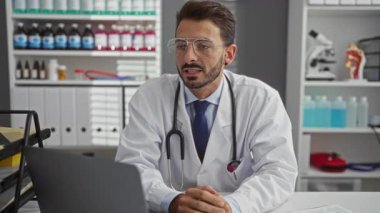 Klinikteki genç İspanyol erkek doktor laptopla danışmanlık yapıyor. Etrafında tıbbi malzemeler ve çizelgeler var..