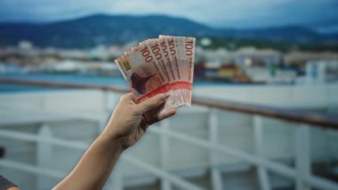 Elinde Norveç kronu olan bir adam, arka planda manzaralı dağlar olan bir yolcu teknesinde seyahat ve finansal temalar sergiliyor..