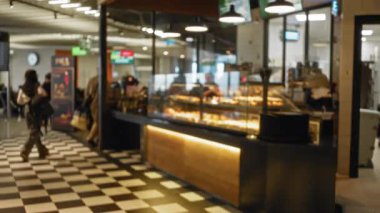 Fırında bokeh efektli bulanık bir kadın, yoğun bir iç ve pasta görüntüsü sergiliyor, dağıtılmış müşteriler ve modern market ortamında ortam aydınlatması sunuyor.