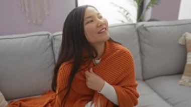 Sıcak turuncu bir battaniyeye sarılmış, rahat bir oturma odasında gülümseyen bir kadın..