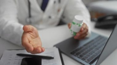 Elinde ilaç şişesi olan bir doktor, bir klinikte dizüstü bilgisayar ve belgelerle masada oturuyor, profesyonel bir sağlık hizmeti sunuyor..