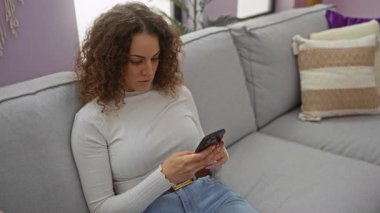 Gri kanepede rahat bir oturma odasında akıllı telefonuyla dinlenen, sakin bir ev ortamı sergileyen bir kadın..