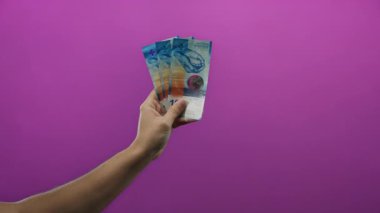 Bir adamın eli İsviçre frangı banknotlarını canlı mor bir arka plana karşı tutuyor, izole bir yaratıcı ortamda para, finans ve ekonomi sergiliyor..