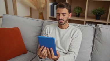 Genç bir adam, rahat bir oturma odasında, bir video görüşmesi sırasında tablet kullanıyor, modern ev teknolojisi ve rahatlık sergiliyor, davetkar bir apartman dairesinde..