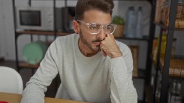 Ofiste oturan genç adam, düşünceli bir ifadeyle modern çalışma odasında gözlük takıyor ve arka planda raflar var..