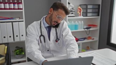 Klinikteki yakışıklı İspanyol erkek doktor. Laptop ve telefonla birden fazla iş yapıyor. Profesyonel ofis ortamında tıbbi ekipmanlarla çevrili..