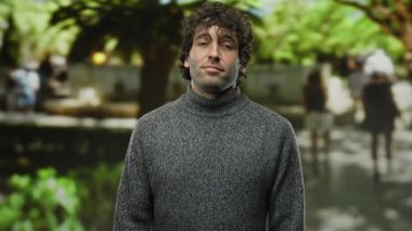 Şehirdeki genç İspanyol adam, bulanık ağaçlar ve insanlarla çevrili gri süveter giyerken silah jesti yapıyor..