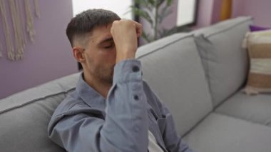 Genç adam rahat bir oturma odasında, modern bir kanepede dinleniyor, rahat bir gömlek giyiyor, düşünceli bir yüz ifadesi var, yumuşak ışıklandırma ve minimalist dekorla çevrili..