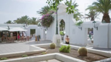 Lanzarote, İspanya 'daki bulanık açık hava sahnesinde beyaz şemsiyeler altında yemek yiyen insanlar ve palmiye ağaçları ve kaktüslerle beyaz bir mimari yapının yanından geçen bir kadın görülüyor.