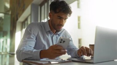 Ofisteki genç adam akıllı telefon ve dizüstü bilgisayar kullanıyor, odaklanmış ve meşgul, masasında kahve fincanı ve belgeler, parlak, modern kapalı bir ortamda.