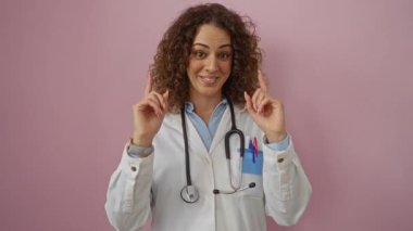 Doktor iki eliyle işaret ediyor ve gülümsüyor, steteskop takıyor, pembe bir arka plana yaslanıyor, kendine güven ve pozitiflik yayıyor..