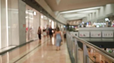 Bulanık insanlar, modern mimari ve mağazaların vitrinlerini gösteren bir arka planla bir alışveriş merkezinde yürüyorlar.