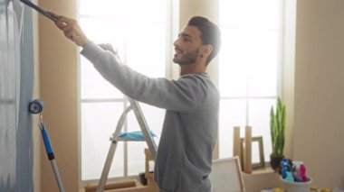 Genç adam yeni evinde duvarları boyuyor, gülümsüyor, silindir kullanıyor, merdivenle ve bitkilerle modern apartmanda..