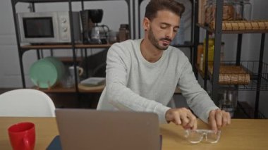 Kapalı ofis ortamında dizüstü bilgisayarda çalışan, gözlük takan, mutfak eşyalarıyla çevrili, ahşap bir masada otururken odaklanmış bir ifade sergileyen genç adam..