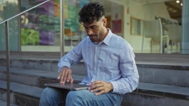 Şehirdeki basamaklarda oturan sakallı genç adam şehir ortamında dizüstü bilgisayar kullanıyor..