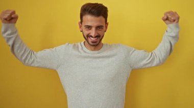 Genç adam kendinden emin bir şekilde gülümsüyor ve parlak sarı bir arka plana karşı kaslarını esnetiyor, açık renk bir kazak giyiyor, güç ve pozitiflik saçıyor..