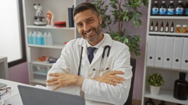 Kolları çapraz kesişen İspanyol erkek doktor tıbbi ekipman ve bitkilerle dolu modern bir hastane kliniğinde güvenle gülümsüyor, profesyonelliği ve sağlık hizmetlerini resmediyor..